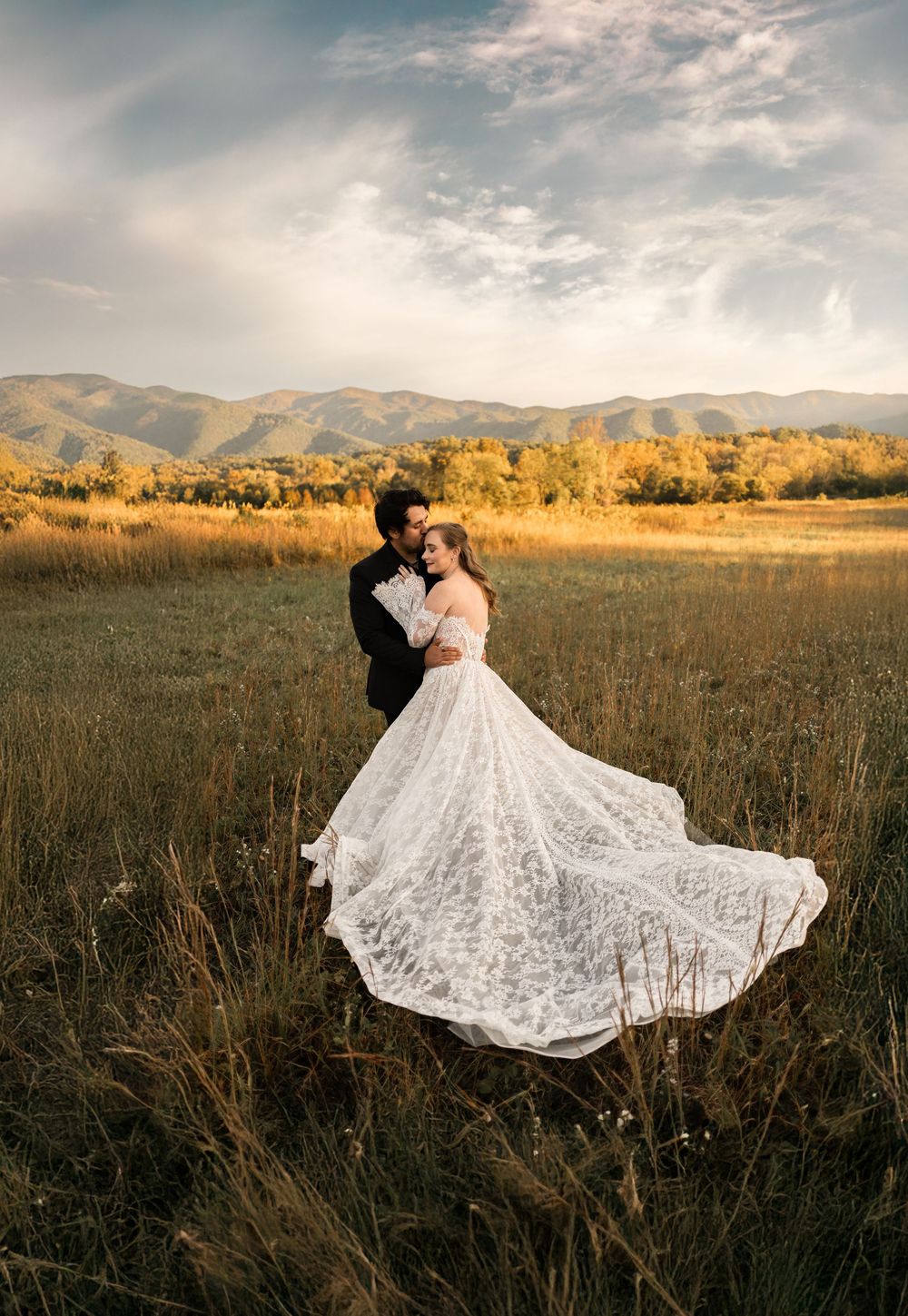 A couple in wedding attire embraces in a golden field at sunset with mountains in the background.