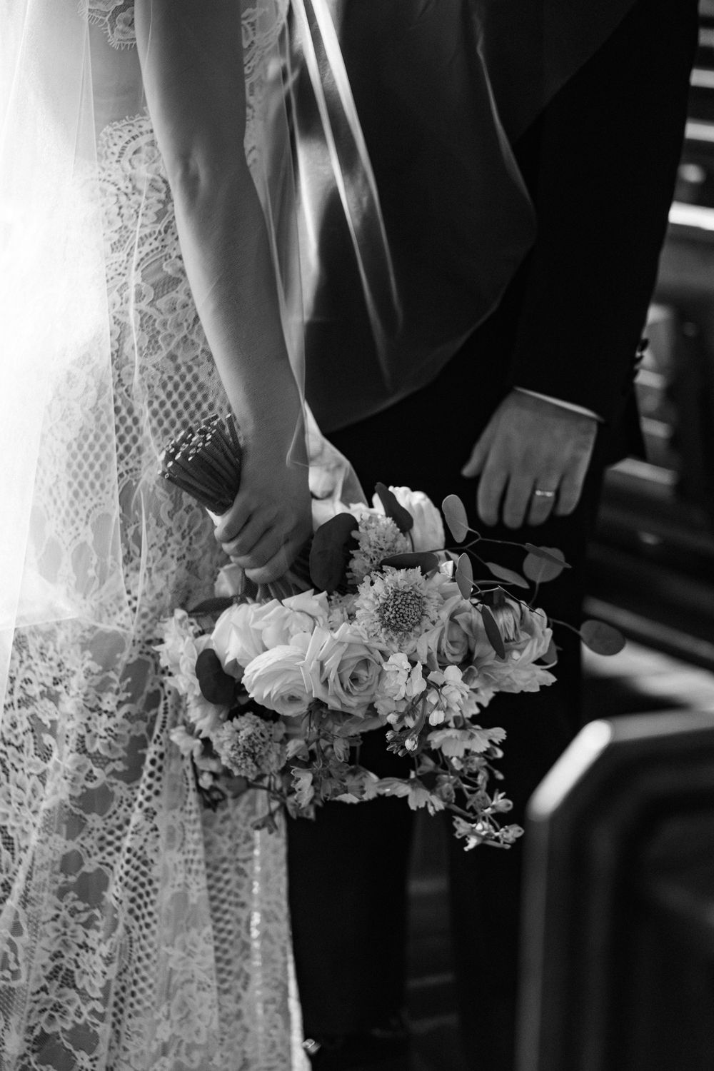 Black and white close-up of a wedding bouquet held against a lace dress with elegant floral details.