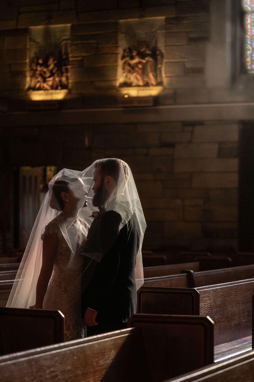 A couple sits close together in a dimly lit church pew with light streaming through stained glass windows.