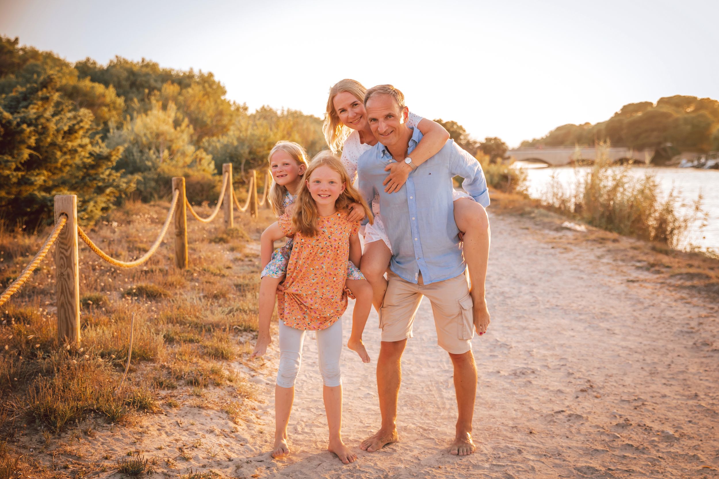 A dad giving a piggyback ride to his wife while walking a scenic path with their two young girls during a family session in Mallorca.