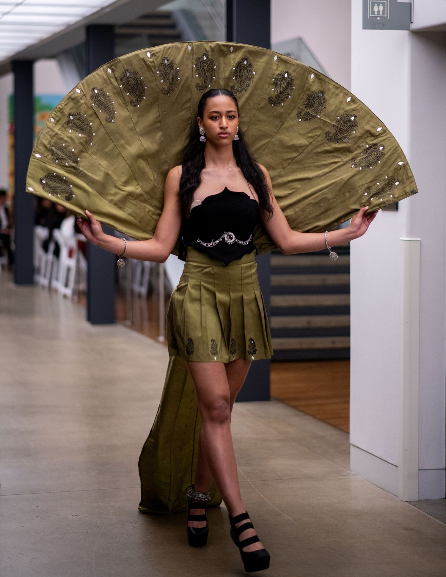 Model in black top and olive skirt poses with dramatic gold fan-shaped accessory on runway.