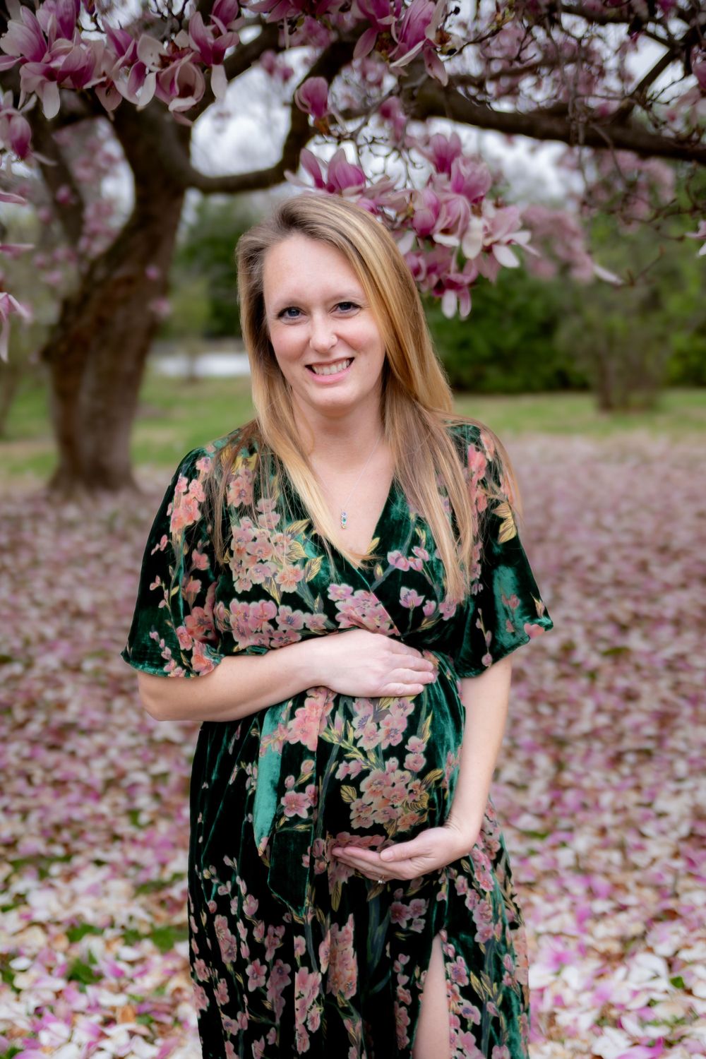 Expectant woman in floral green dress poses under blooming pink magnolia tree with petals scattered on ground.