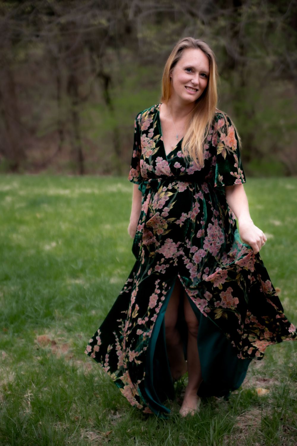 A person wearing a flowing black floral maxi dress poses outdoors on green grass in a springtime setting.