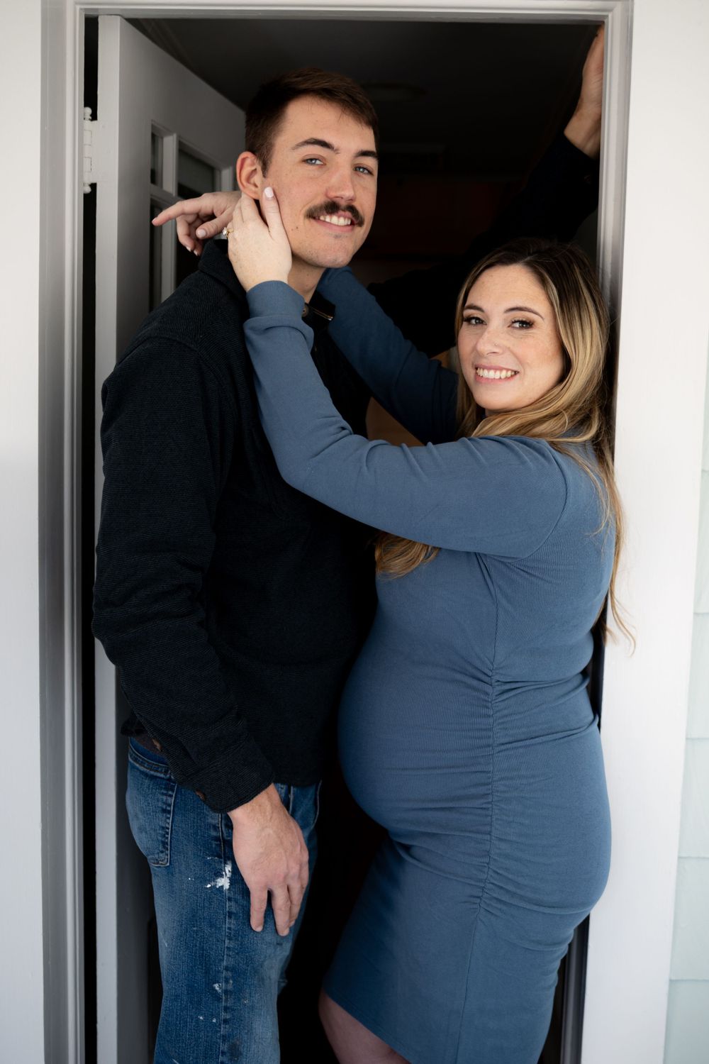A couple standing in a doorway, with the woman in a blue fitted dress touching the man's face in a tender moment.
