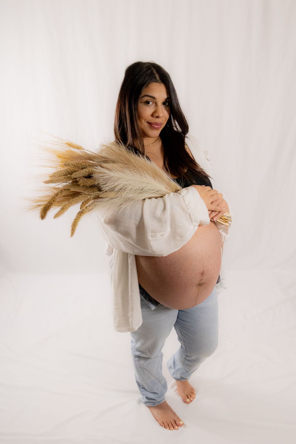Expectant mother in white top and jeans poses with dried pampas grass against white backdrop in maternity photo.
