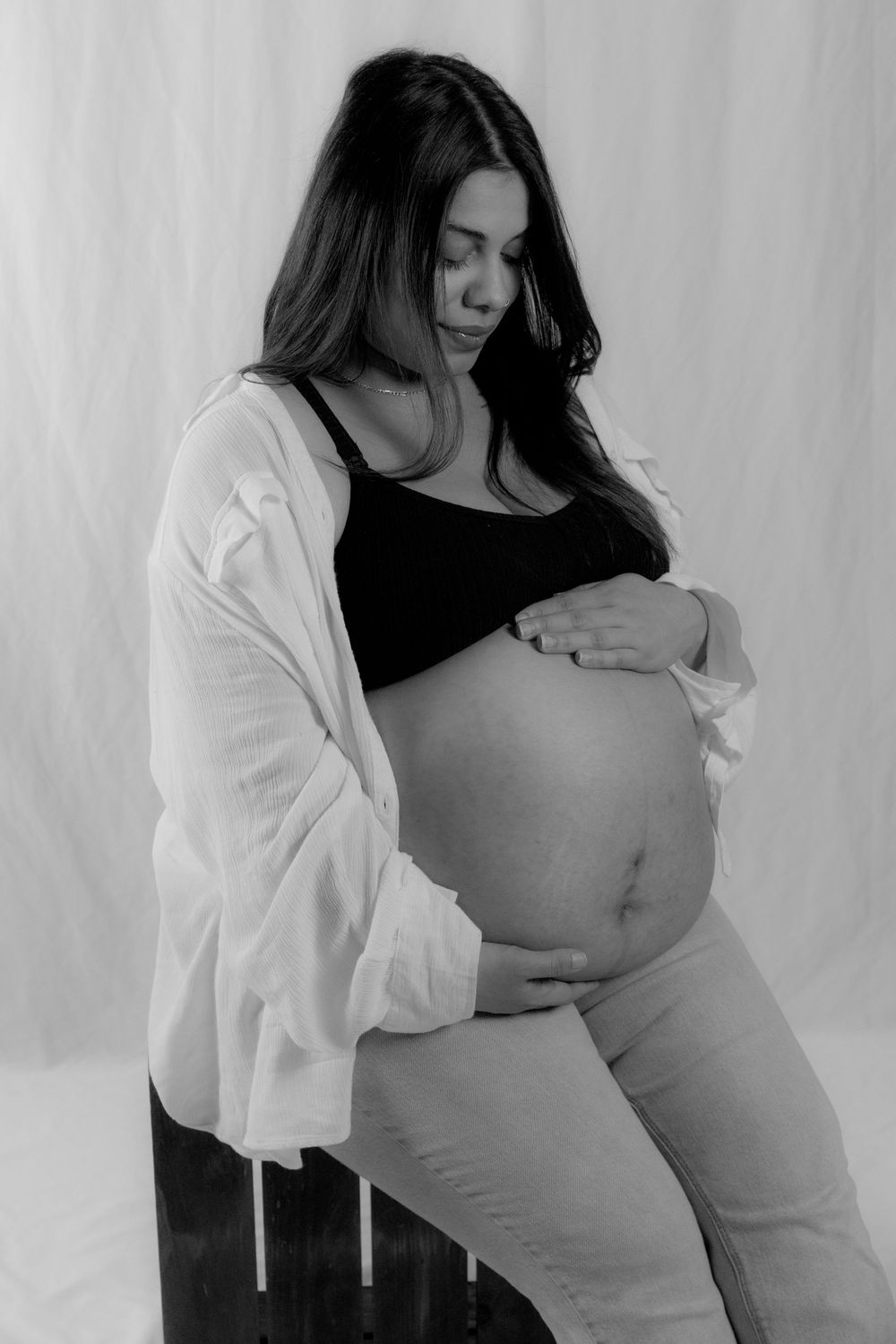 A black and white maternity photo shows an expectant mother in a white cardigan and black top cradling her pregnant belly.