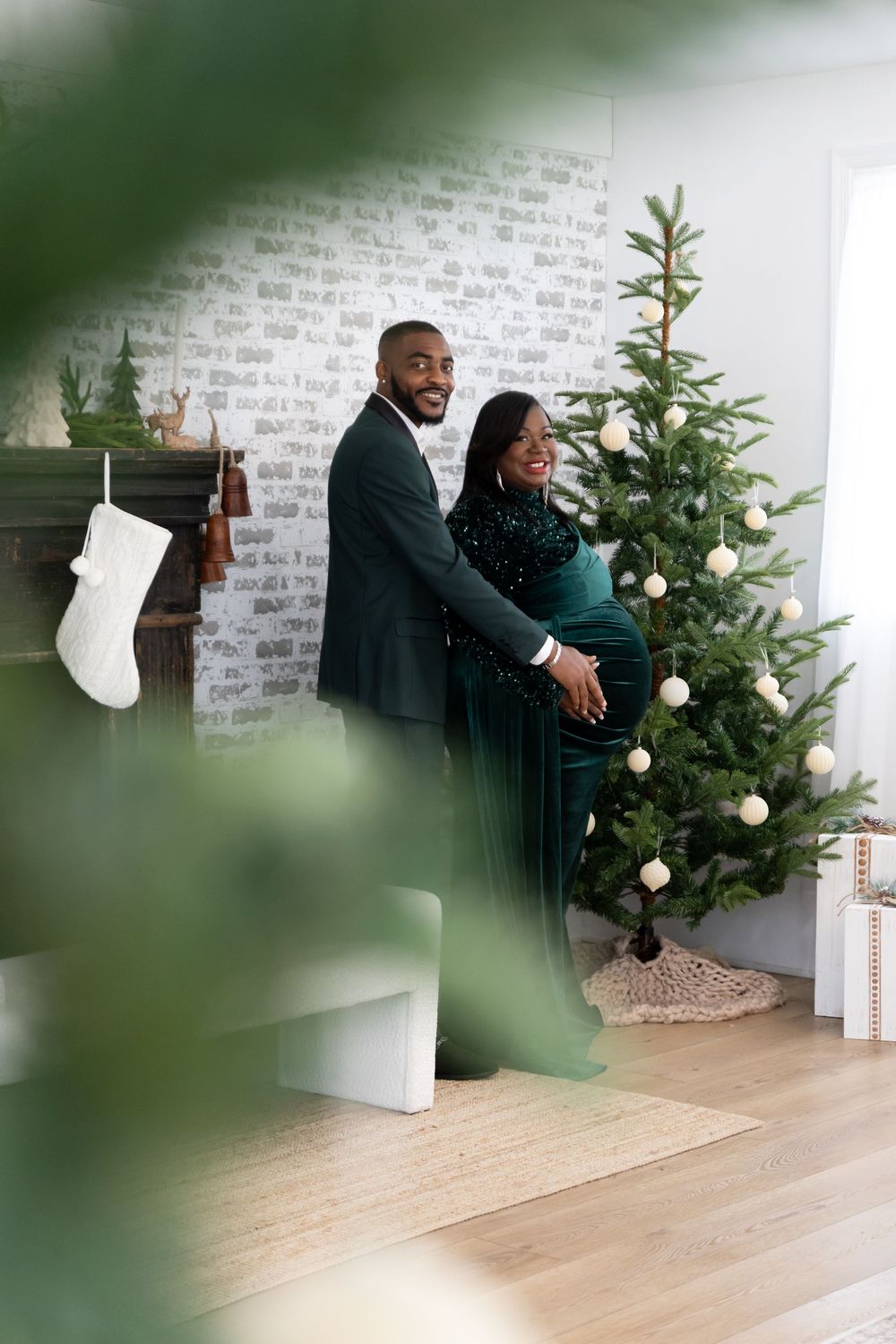 A couple sits together on the floor beside a decorated Christmas tree in a modern minimalist living room setting.