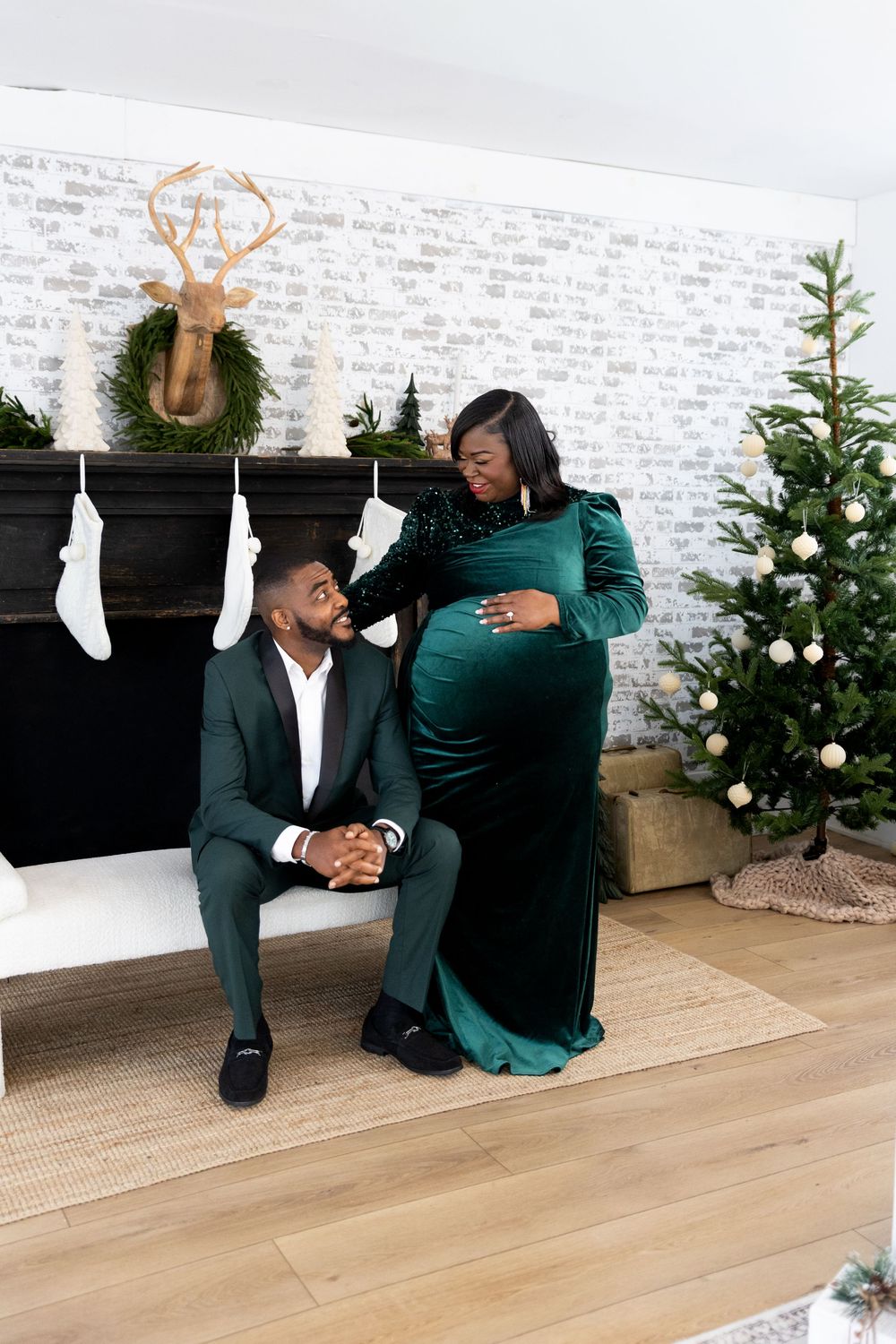 A couple in formal attire shares a sweet proposal moment in front of a decorated Christmas tree and white brick wall.