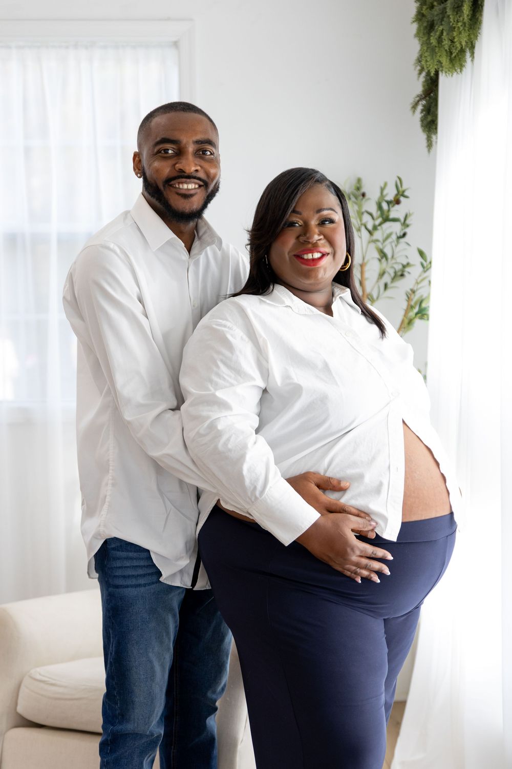 A couple in white tops and dark pants embrace lovingly in a bright room with white curtains and greenery.