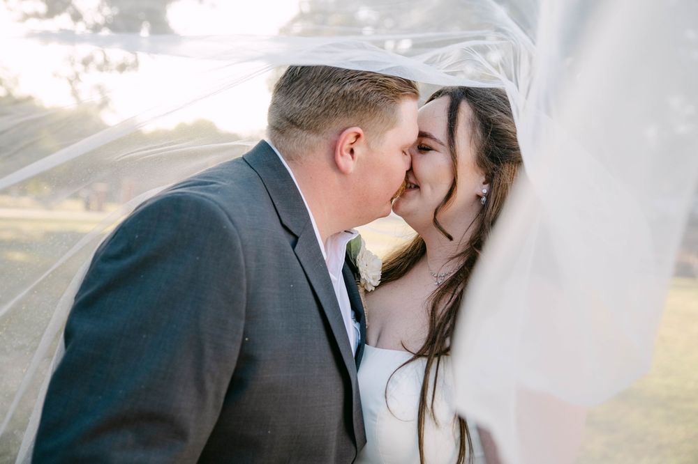 A romantic wedding couple shares a tender kiss under a flowing white veil during their outdoor ceremony.
