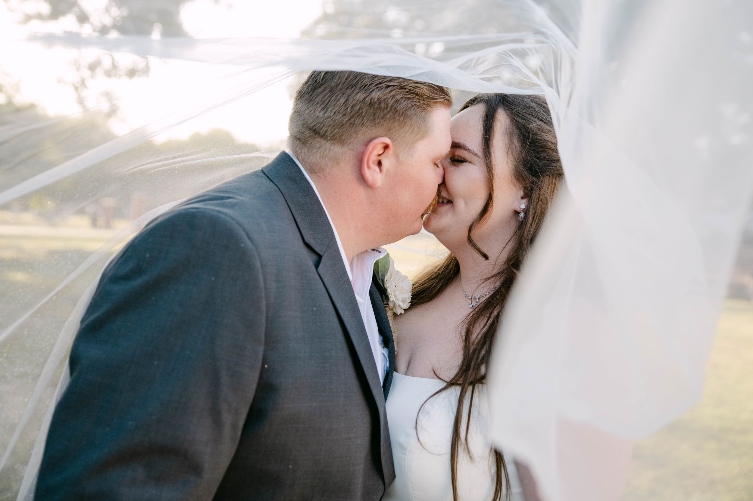 A romantic wedding couple shares a tender kiss under a flowing white veil during their outdoor ceremony.