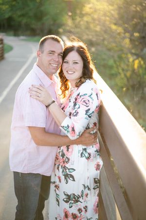 A couple poses together on a path during a sunset photoshoot, with a floral dress and pink shirt glowing in golden light.