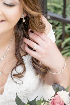 Close-up detail of bridal jewelry with delicate pearl necklace and earrings against white lace dress and pink roses.