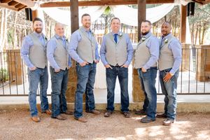 Groomsmen wearing matching gray vests and blue jeans pose together under wooden beams at rustic outdoor venue.