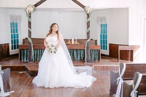 A bride in a flowing white wedding gown poses gracefully in a rustic church venue with hardwood floors and white walls.