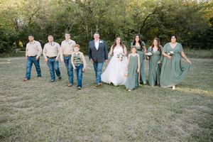 Wedding party in coordinated blue and casual attire poses together on grassy field during sunset.