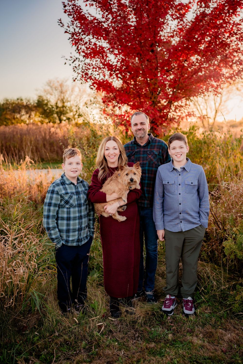 A family with their goldendoodle dog poses in front of a vibrant red maple tree during golden hour.