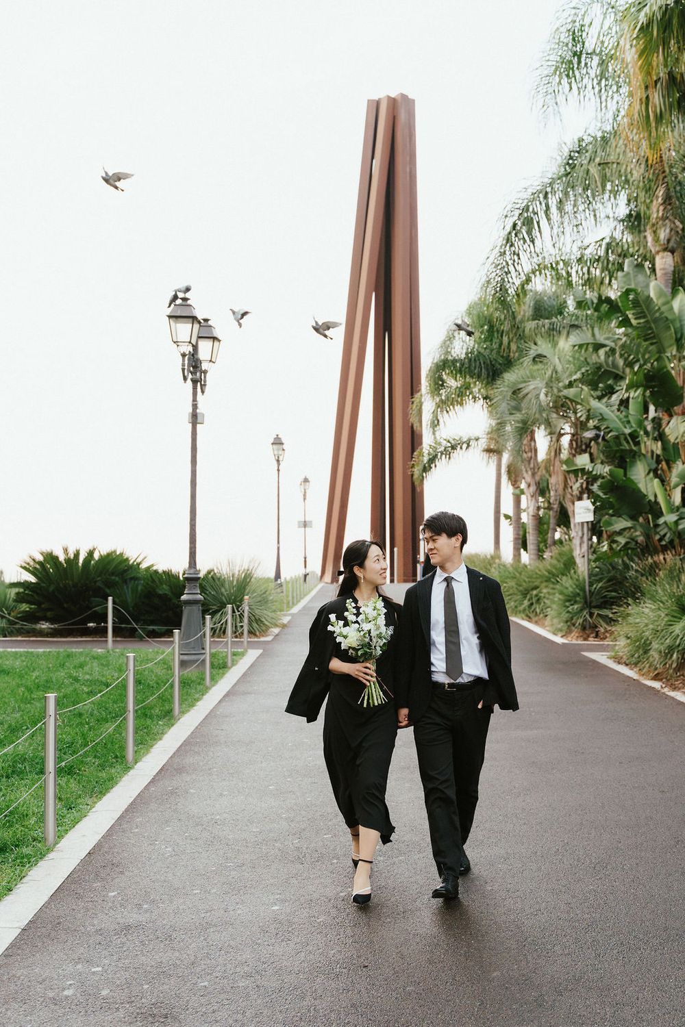 Couple in black suits walking along a palm-lined path, woman holding white flowers, birds flying overhead.