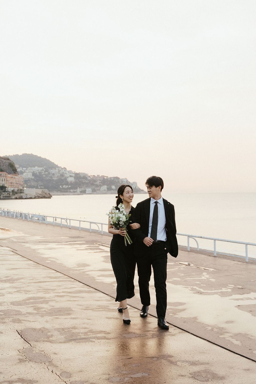 Couple in black formal attire walking along a waterfront promenade, woman holding white flowers.