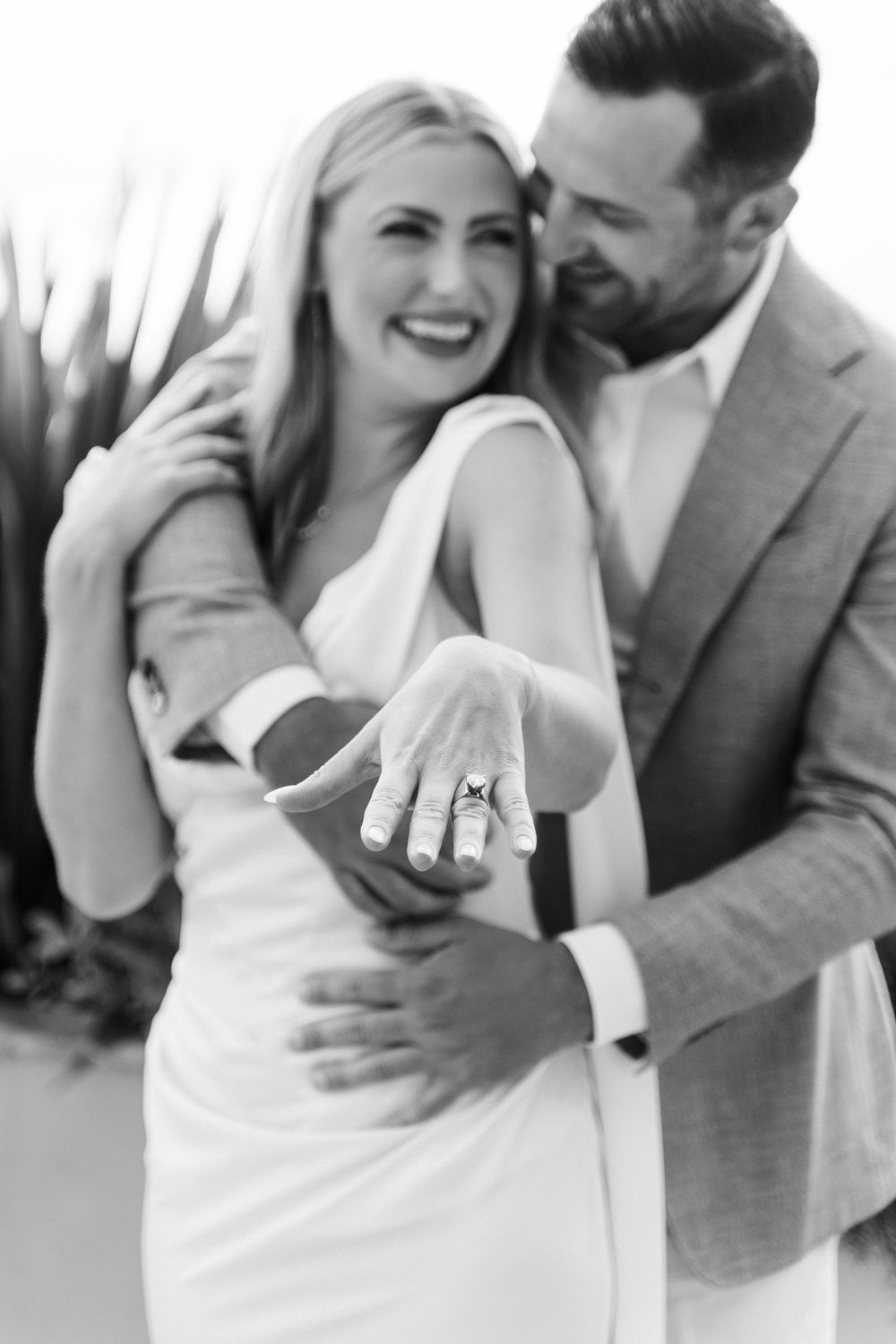 Black and white photo of smiling blonde woman showing off engagement ring while man in suit embraces her from behind.