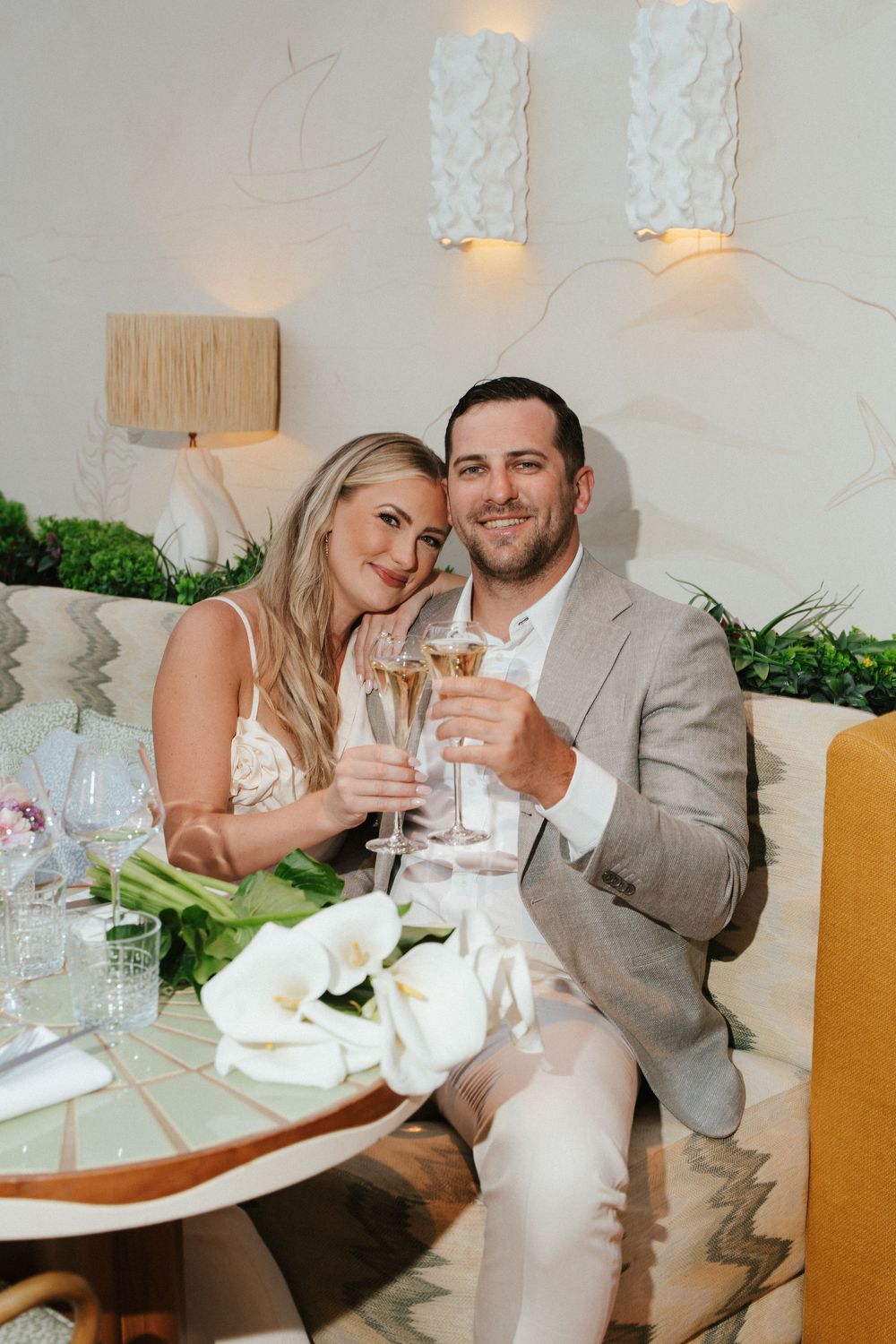 Couple toasting with champagne glasses at elegant restaurant table with white calla lilies, dressed in wedding attire.