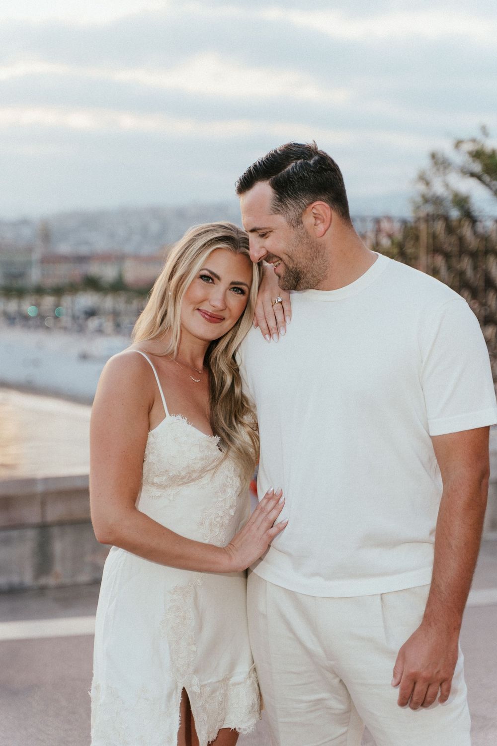 Couple in matching white outfits posing together near a waterfront, woman in lace dress leaning on mans shoulder.