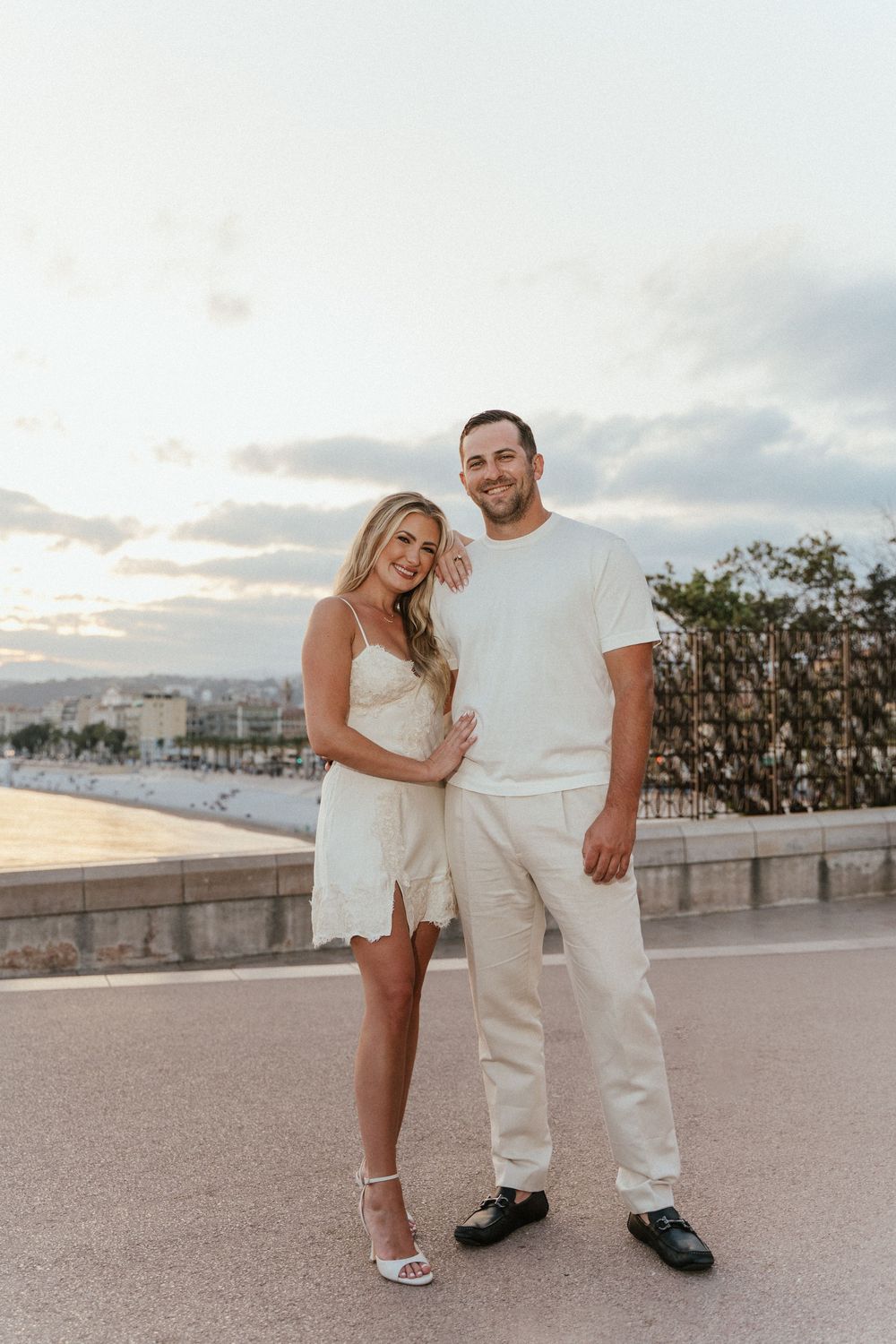 Couple in matching white outfits posing outdoors at sunset near a coastal promenade.