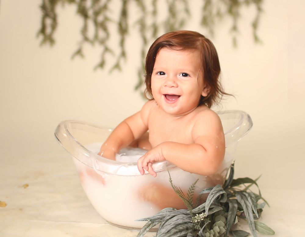 A happy baby sits in a white bowl surrounded by greenery against a light backdrop during a professional photo session.