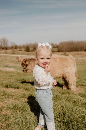 A joyful toddler in light denim and lace stands in a grassy field with a large brown dog.
