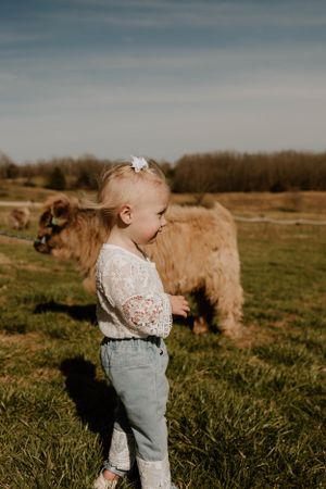 A toddler in a lace top and jeans stands in profile with a brown dog grazing in the background.