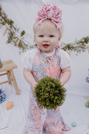 A child wearing a floral outfit and bow holds a green pompom against a white backdrop with greenery.