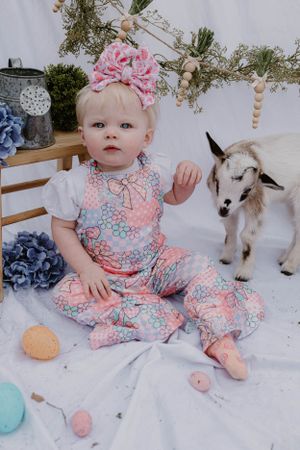 Sweet spring photo setup with pink floral outfit and baby goat companion on white backdrop with Easter eggs.