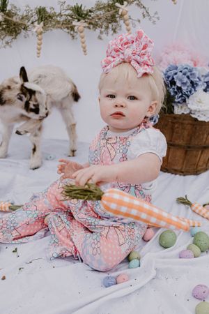 Festive spring scene with floral outfit and baby goat among pastel Easter eggs and decorative elements.