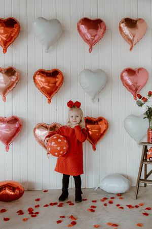 Valentine's Day photo setup with red dress against white wall decorated with pink and red heart balloons and rose petals.