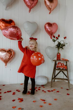 Joyful Valentine's Day scene with heart balloons and red dress against white paneled wall.
