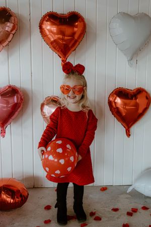 Holiday photoshoot with red heart balloons, red dress, and heart sunglasses against white backdrop.