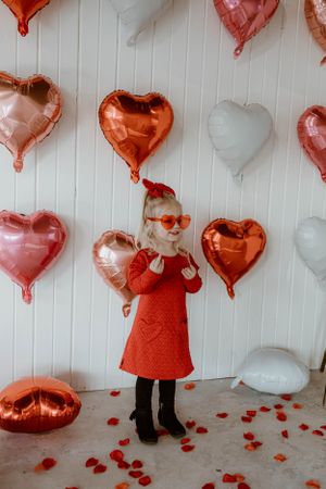 Heart balloon Valentine's setup with orange sunglasses and red dress against white wall.