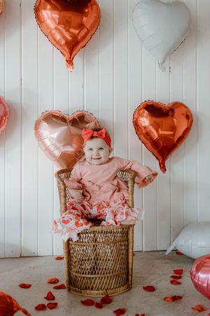 Sweet Valentine's scene with pink outfit in wicker chair surrounded by heart balloons.