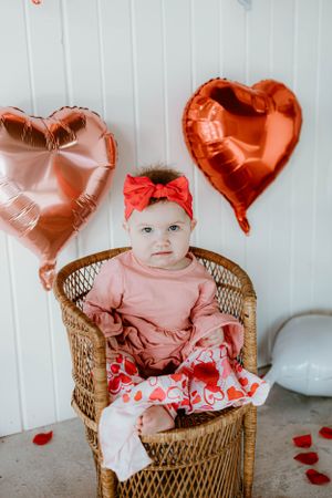 Valentine's Day photo setup with pink dress, wicker chair and rose gold heart balloons against white background.