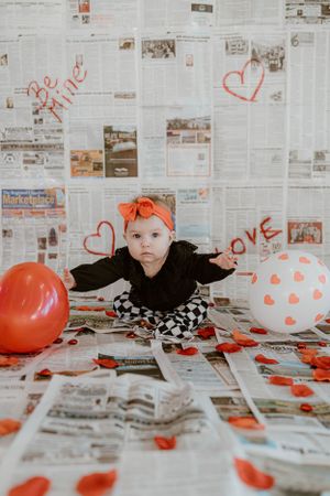 Baby in black outfit with orange headband sits on newspaper backdrop with Valentine's decorations and balloons.