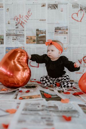 Baby reaches for heart balloon while sitting on newspaper-covered floor with Valentine's decorations.
