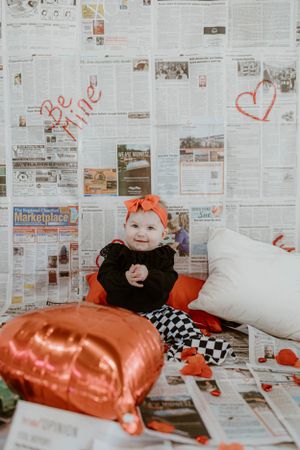 Baby in black outfit with checkered pants sits among Valentine's decorations and heart pillows.