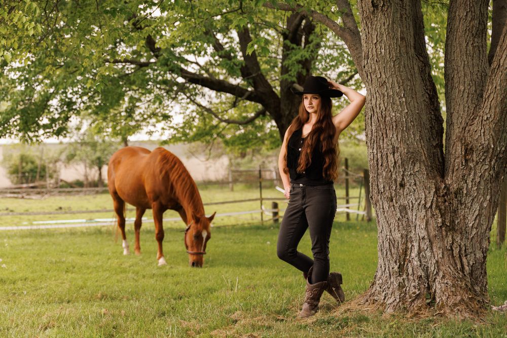 Western-style portrait with chestnut horse grazing near large tree in pastoral setting.