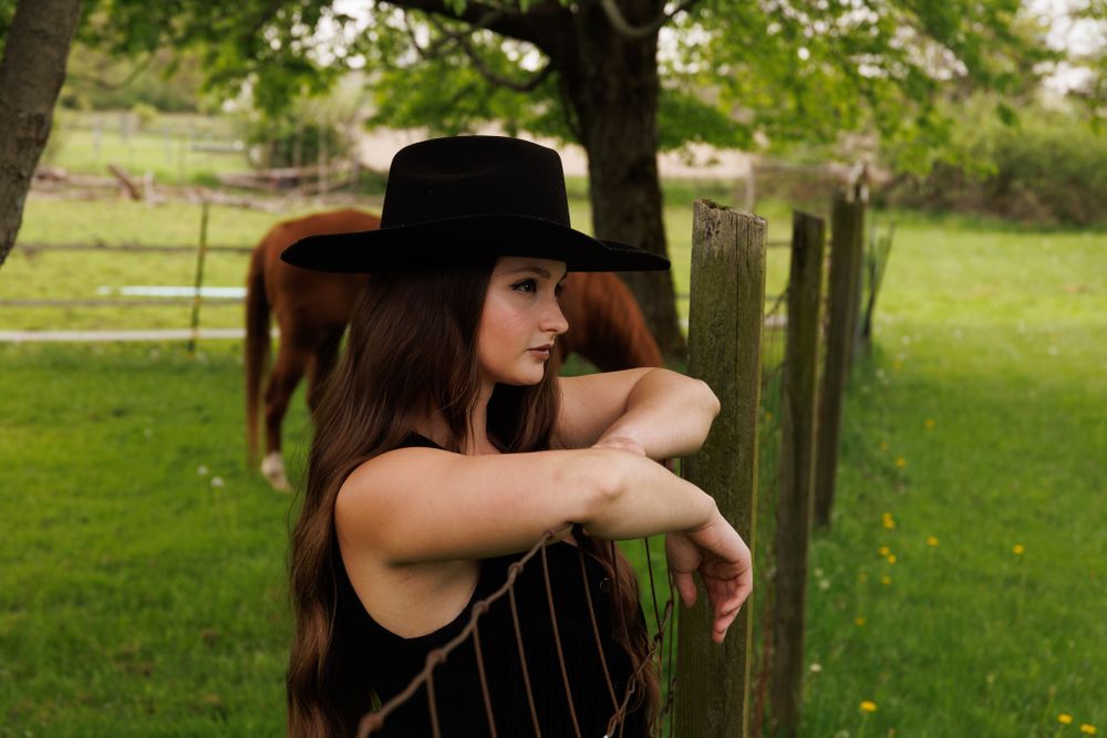 Artistic western portrait against rustic fence with horse visible in background.