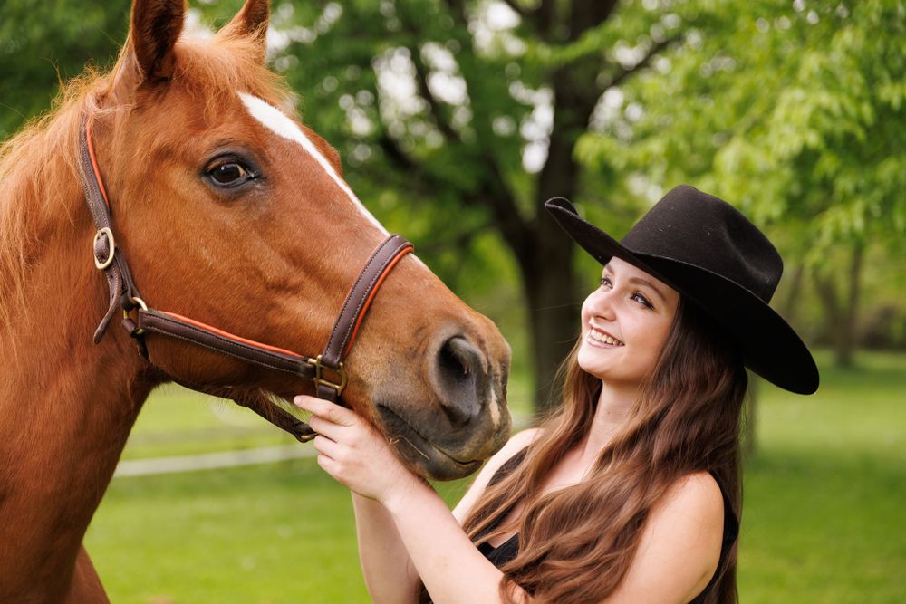 Tender moment between chestnut horse and rider in western hat among green trees.