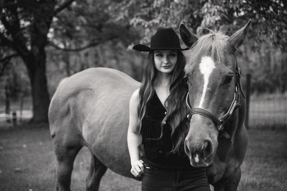 Black and white portrait of a horse and rider standing together in a rustic outdoor setting.