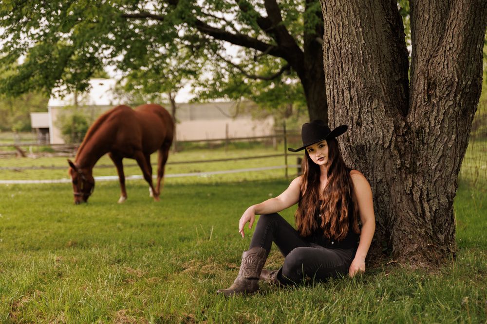 Rustic country scene with horse grazing while rider rests under large shade tree.
