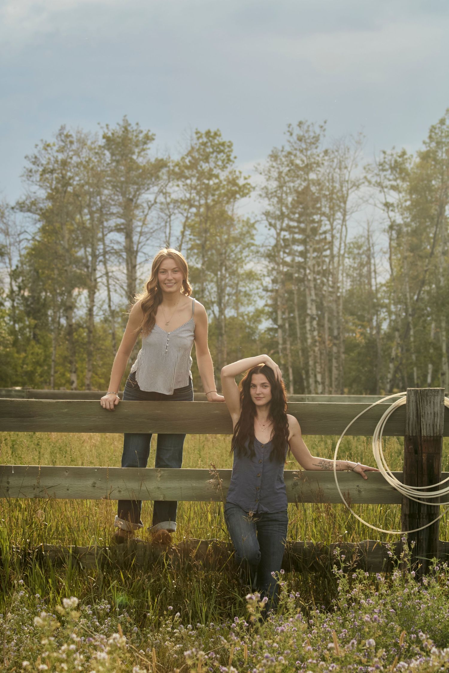 Two young women sitting on a wooden fence in jeans and tank tops, smiling in a rural field with flowers and a rope, summer countryside vibe.