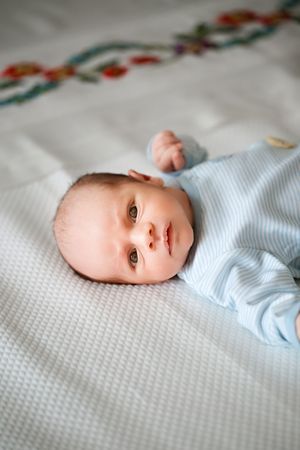Alert newborn baby in light blue sleeper lying on white textured bedding.