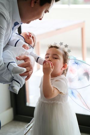 A young child in a white tulle dress interacts with a baby while standing near a window.